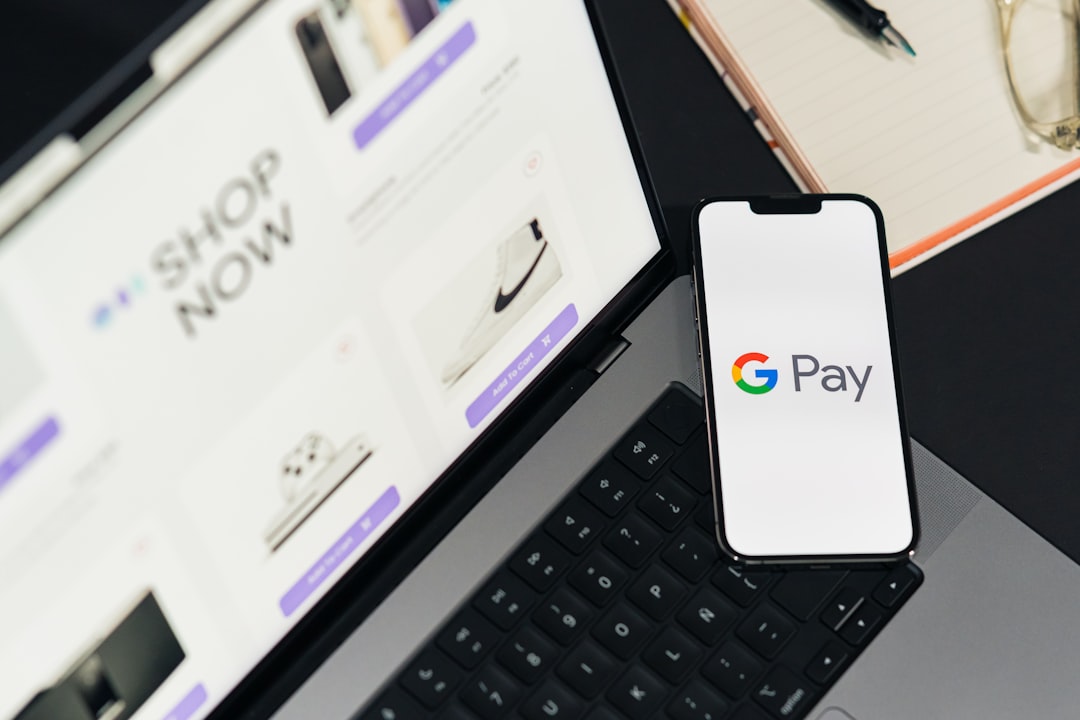 Google Pay on phone screen. Top view of workspace. Table desk with laptop with ecommerce on screen, mobile, notepad